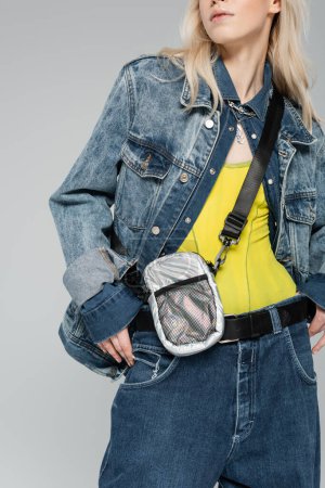 cropped view of blonde woman in stylish denim jacket with belt bag posing isolated on grey 