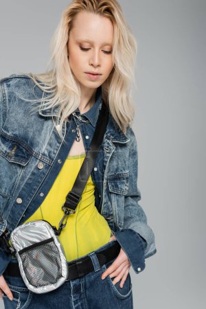Téléchargez les photos : Albino blonde woman in stylish denim jacket with belt bag looking down while posing isolated on grey - en image libre de droit
