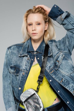 young woman in stylish denim jacket posing while adjusting blonde hair isolated on grey 