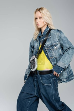 young blonde woman in stylish denim jacket posing with belt bag isolated on grey 