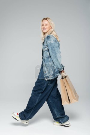 Téléchargez les photos : Pleine longueur de jeune femme heureuse en tenue de denim élégant tenant des sacs à provisions et marchant sur gris - en image libre de droit