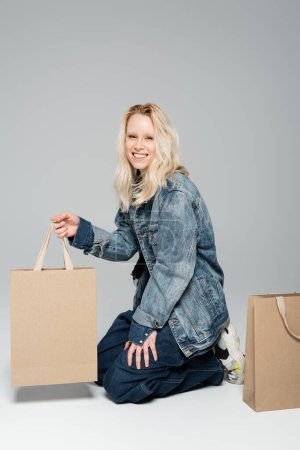 joyful young woman in stylish denim outfit sitting around shopping bags on grey 
