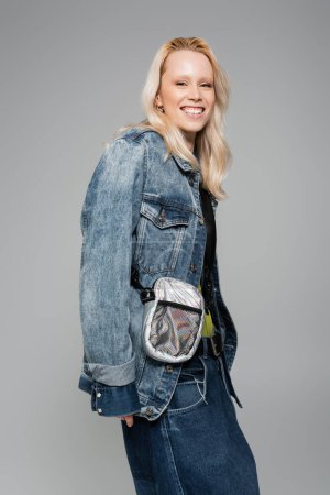 Foto de Cheerful blonde woman in denim blue jacket and belt bag smiling isolated on grey - Imagen libre de derechos
