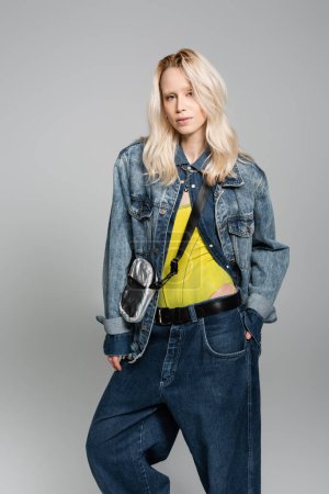 Téléchargez les photos : Young blonde woman in denim blue jacket and belt bag posing with hand in pocket isolated on grey - en image libre de droit