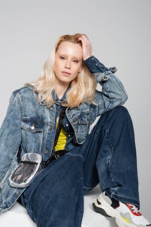 Foto de Blonde woman in blue denim outfit and trendy jeans sitting on white cube isolated on grey - Imagen libre de derechos