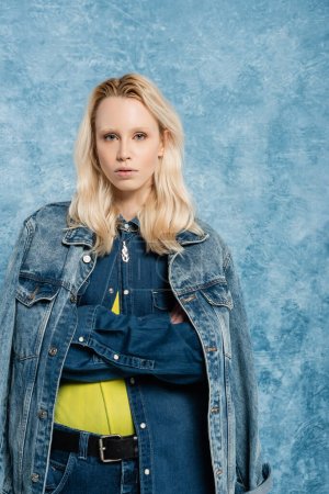 blonde woman in denim jacket posing with crossed arms near blue textured background  