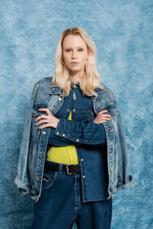blonde model in denim jacket posing with crossed arms near blue textured background  