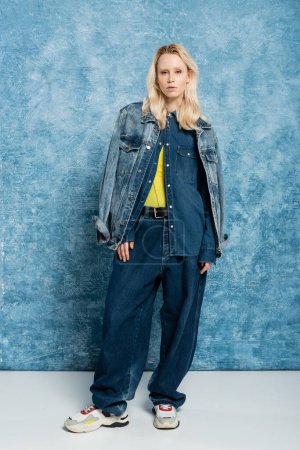 full length of blonde woman in denim outfit posing near blue textured background  
