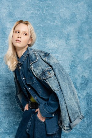 blonde woman in denim jacket posing with hands in pockets near blue textured background  