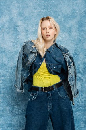 blonde woman in denim jacket and trendy jeans posing near blue textured background  