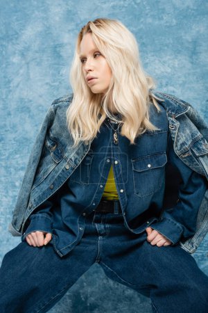 Blondine in Jeansjacke posiert und schaut in der Nähe von blauem Hintergrund weg  