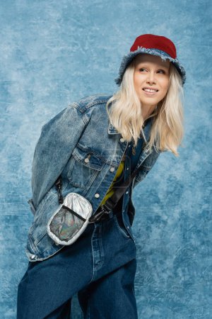 happy blonde woman in denim jacket and panama hat posing near blue textured background  