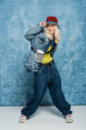 full length of happy blonde woman in denim outfit adjusting panama hat near blue textured background  