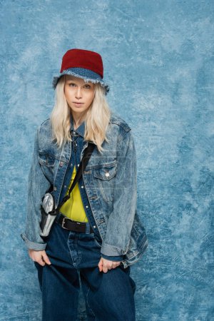 blonde woman in denim jacket and panama hat adjusting trendy jeans near blue textured background  