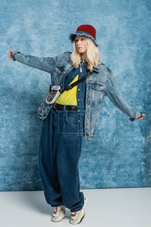 full length of blonde woman in denim jacket and panama hat posing with outstretched hands near blue textured background  