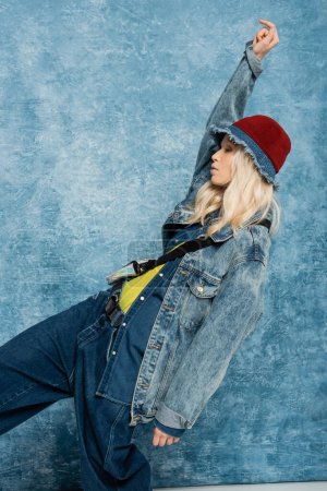 blonde woman in denim jacket and panama hat looking away and posing with raised hand near blue textured background  