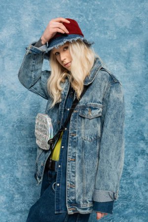 blonde woman in denim jacket and panama hat looking at camera near blue textured background  