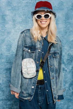 glückliche blonde Frau im Jeans-Outfit posiert mit Panamahut und Sonnenbrille vor blauem Hintergrund  