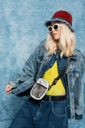 blonde woman panama hat and sunglasses adjusting denim jacket near blue textured background  