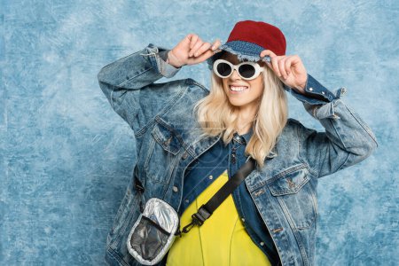 smiling blonde woman in denim jacket and sunglasses adjusting panama hat near blue textured background  