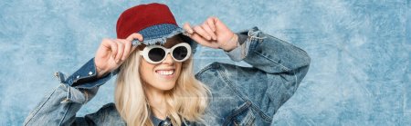smiling blonde woman in denim jacket and sunglasses adjusting panama hat near blue textured background, banner 