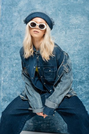 young blonde woman in denim outfit posing in panama hat and sunglasses near blue textured background  
