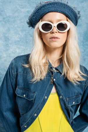 young blonde woman in denim panama hat and sunglasses posing near blue textured background  