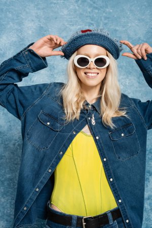 positive blonde Frau mit Jeans-Panamahut und Sonnenbrille posiert in der Nähe von blau strukturierten Hintergrund  