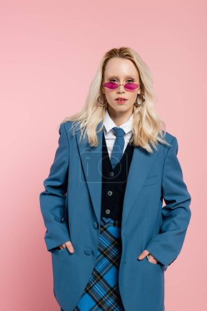 blonde woman in blue blazer with tie and sunglasses posing with hands in pockets isolated on pink 