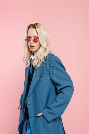 young blonde woman in blue blazer with tie and sunglasses posing with hands in pockets isolated on pink 
