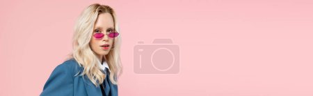 young blonde woman in blue blazer with tie and sunglasses posing with hands in pockets isolated on pink, banner 