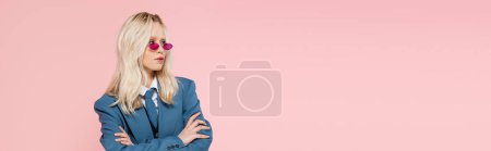 blonde woman in blue blazer with tie and sunglasses posing with crossed arms isolated on pink, banner 