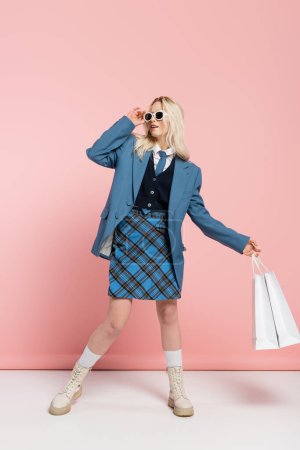 full length of blonde woman in blue outfit adjusting trendy sunglasses and holding shopping bags on pink 