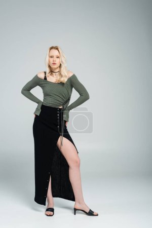 Téléchargez les photos : Pleine longueur de jeune femme blonde à manches longues et sexy jupe noire posant avec les mains sur les hanches sur le gris - en image libre de droit