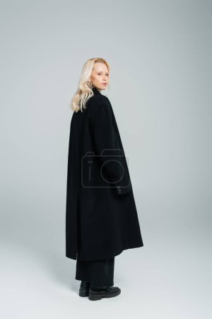 Téléchargez les photos : Pleine longueur de jeune femme blonde en manteau noir debout sur le gris - en image libre de droit