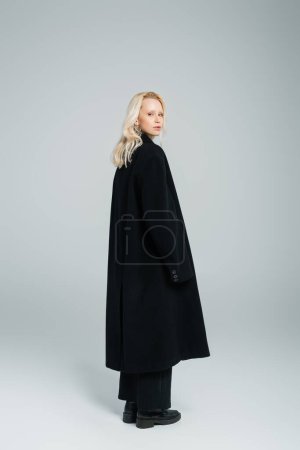 Foto de Longitud completa de la joven rubia en abrigo negro de pie y mirando a la cámara en gris - Imagen libre de derechos