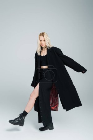 Téléchargez les photos : Pleine longueur de femme blonde en manteau noir et jupe regardant la caméra tout en posant sur le gris - en image libre de droit