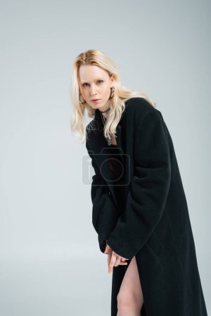 blonde woman in black skirt and coat posing while looking at camera isolated on grey 