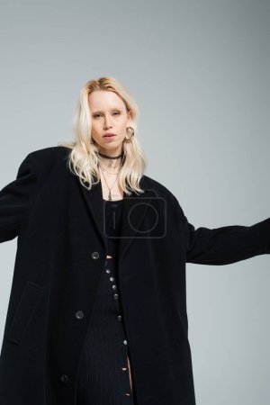 Téléchargez les photos : Jeune femme blonde en manteau noir tendance regardant la caméra isolée sur gris - en image libre de droit