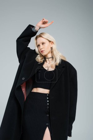 young blonde woman in trendy black coat looking away and gesturing isolated on grey 