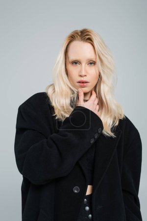 Téléchargez les photos : Blonde and young woman in stylish black coat looking at camera isolated on grey - en image libre de droit