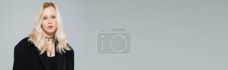 Téléchargez les photos : Blonde et jeune femme en manteau noir élégant regardant la caméra tout en se tenant isolé sur gris, bannière - en image libre de droit