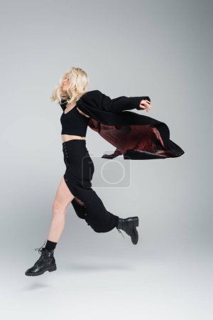 Téléchargez les photos : Full length of blonde and young woman in stylish black outfit jumping on grey - en image libre de droit