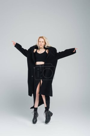 full length of blonde and young woman in stylish black outfit levitating on grey 