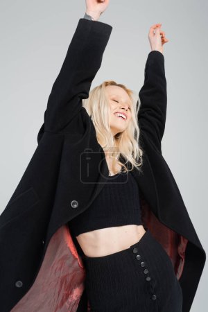 Foto de Excited young woman in stylish black outfit posing with raised hands isolated on grey - Imagen libre de derechos