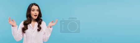 Téléchargez les photos : Surprised young woman in sleepwear gesturing while standing with opened mouth isolated on blue, banner - en image libre de droit