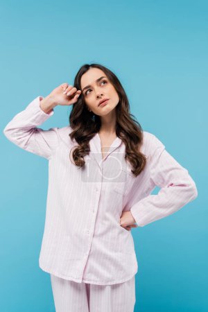 pensive young woman in sleepwear looking away and standing with hand on hip isolated on blue 