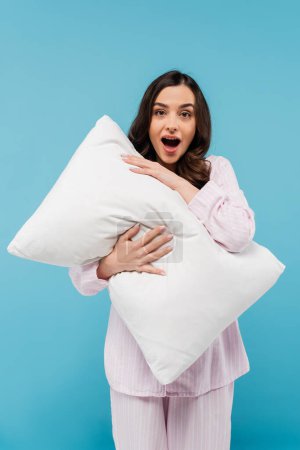 Schockierte junge Frau in Schlafanzügen hält weißes Kissen auf blauem Grund 