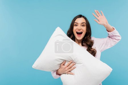 verblüffte junge Frau in Schlafanzügen hält weißes Kissen in der Hand und winkt vereinzelt auf blauem Grund 