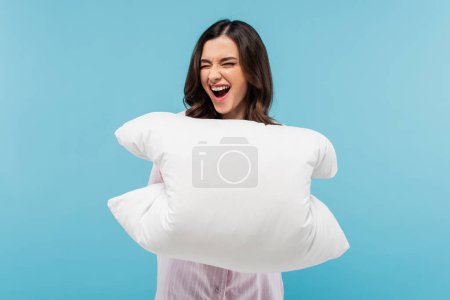 aufgeregte junge Frau in Schlafanzügen hält weißes Kissen auf blauem Grund 